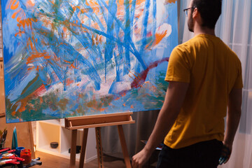 Male artist steps back from a large blue and orange abstract canvas to inspect the composition and evaluate progress during the creative process. © DC Studio