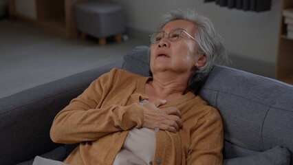 Fototapeta premium Senior woman reclines on a gray sofa with one hand on her chest, looking upward and breathing through visible discomfort while experiencing sudden upper body pain alone inside her own home.