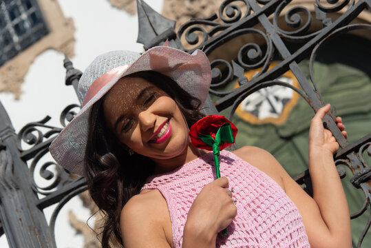 A smiling young Brazilian woman in a pink crochet dress and a red flower, standing in front of an old gate.
