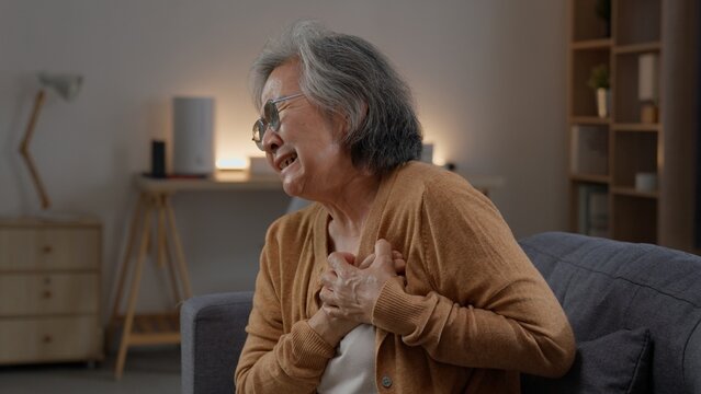Senior woman with glasses sits on a sofa clutching her chest with both hands, showing visible discomfort and distress while experiencing sudden upper body pain alone in a quiet living room.