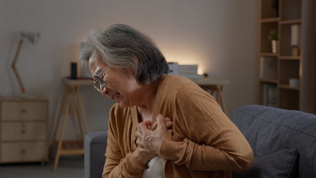 Elderly woman bends forward on a sofa while pressing both hands to her chest, grimacing with intense discomfort during a sudden episode of upper body pain while staying alone at home today.