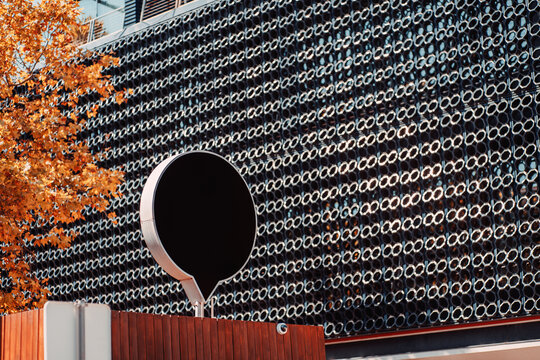 Advertising mockup featuring a blank black circular billboard mounted before a patterned metal building facade, framed by warm autumn foliage for bold brand presentation