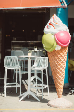 Selective focus on a small outdoor cafe table and white chairs beside a giant colorful ice cream cone display, creating a playful setting for summer hospitality concepts