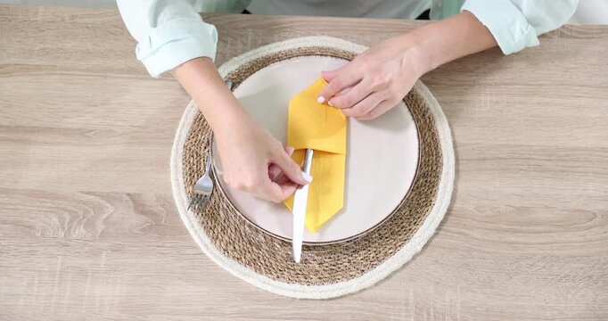 Woman folding yellow napkin and putting cutlery into it at wooden table indoors, above view. Camera moving in