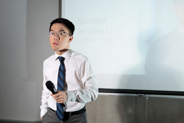 A young Asian man in a white shirt and tie stands beside a projected screen while holding a microphone, for public speaking, business training, or conference communication. © fotolite