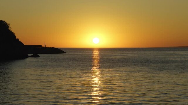 Beautiful orange sun slowly descending over the calm sea with a rocky coast silhouetted in Marseille