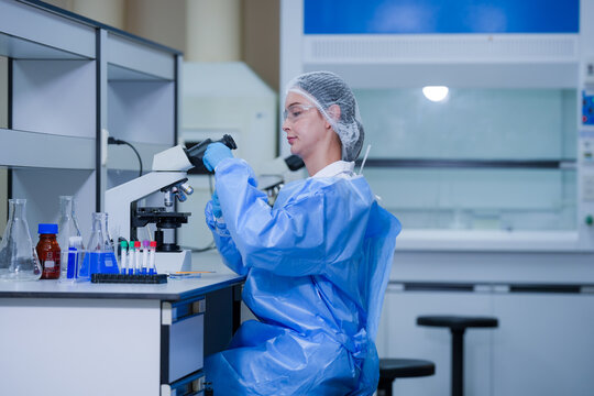 A female microbiologist in a white coat, protective eyewear and a sterile hairnet raising a tube beside the microscope inside a university biotechnology laboratory.