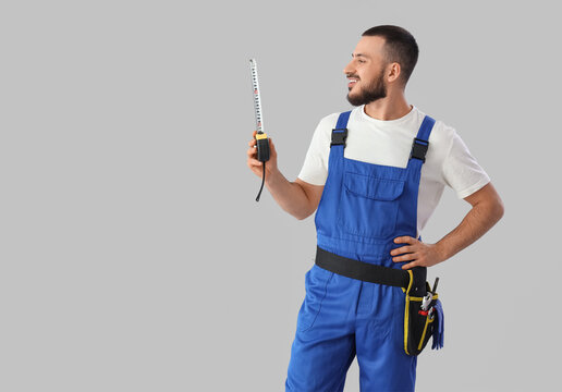 Young male construction worker with measuring tape on grey background