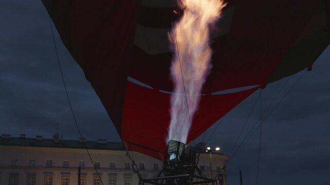 Colorful hot air balloon's burner ignites flames, lighting up the balloon fabric as it prepares for flight in an urban evening setting