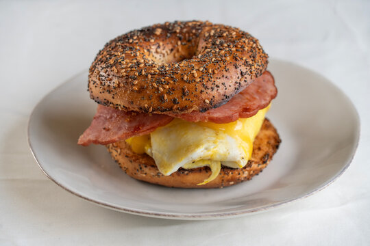 An egg, ham and cheese sandwich on a toasted everything bagel on a white plate.