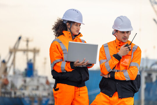 Engineers in high visibility safety gear with laptop and walkie talkie at port, Professional logistics workers reviewing data on computer near cargo ships
