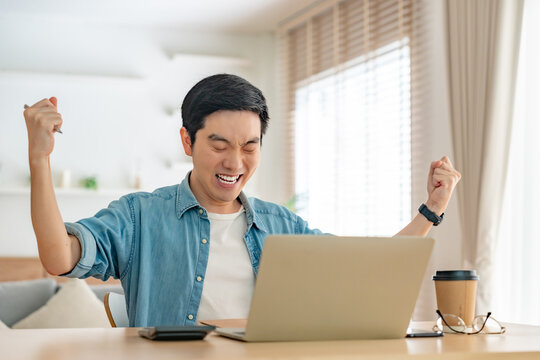 Young Asian excited man freelancer using laptop device working in house. Handsome guy student chatting and typing on computer notebook to communicate online with successful in living room at home
