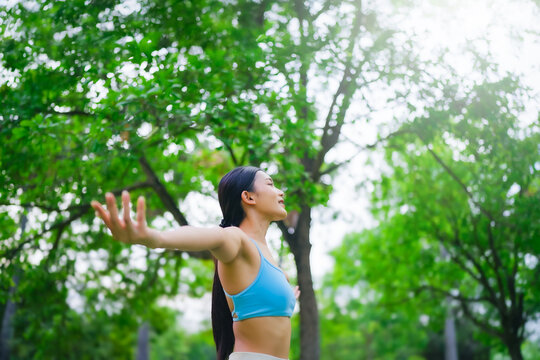 Young Asian woman relaxed breathing fresh air with trees in the background on a sunny day. Concept of wellness, healthy lifestyle, mindfulness, and outdoor fitness