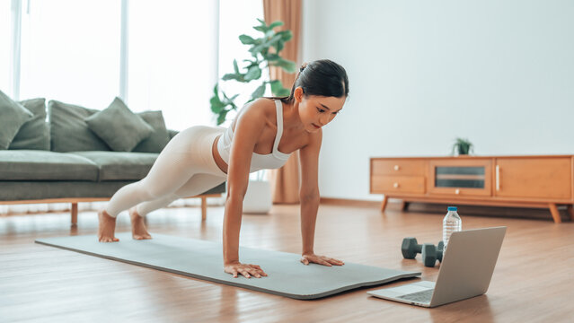 Young Asian woman in sportswear doing plank on training mat while watching online workout tutorials on laptop in the living room. Sport and recreation concept