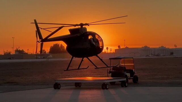 A small helicopter, viewed in silhouette by the setting sun, hovers above and then settles on a landing pad.