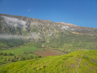 Morning fog drifts along a rugged rocky mountain ridge above a green hillside, with a cultivated field and scattered trees in the valley below. 📍 MahmoudAbad, Sarvabad, Kurdistan, © Hawraman83