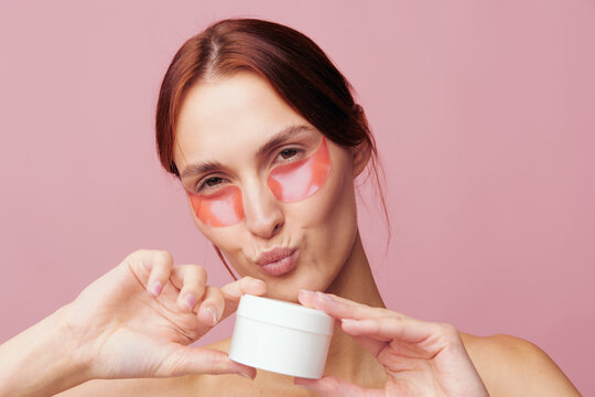 Young woman with pink eye patches holds a white cream jar and pouts her lips. Studio shot against a clean background. Skincare concept for cosmetics, beauty, and wellness.