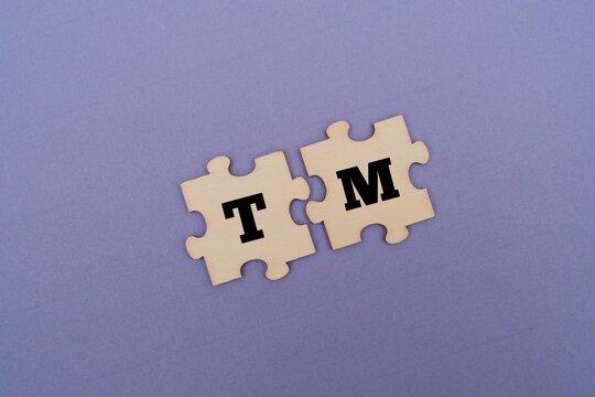 Two interlocking wooden puzzle pieces featuring the letters TM on a coloured background. TM acronym for TOTAL MASTECTOMY 
