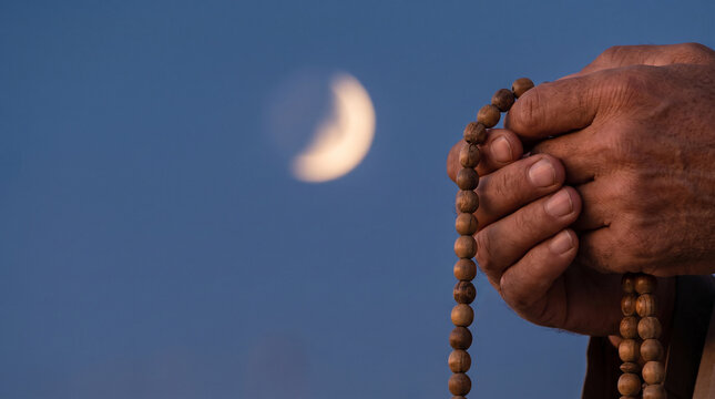 Hands hold wooden prayer beads against a deep blue evening sky featuring a blurred crescent moon.
