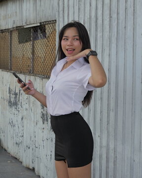 A young Asian woman in a student uniform holds a smartphone by a corrugated metal wall