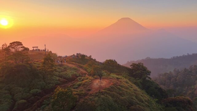 Scenic aerial view of Bukit Sikunir during a golden sunrise, showing lush green hills, misty valleys, and the majestic silhouette of Mount Sindoro in Dieng Plateau, Wonosobo, Indonesia.