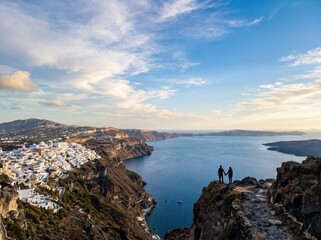 Fototapeta premium A cinematic vertical shot capturing two tiny silhouettes standing on a Santorini cliff during a golden sunrise overlooking the vast Aegean Sea and white miniature architecture.