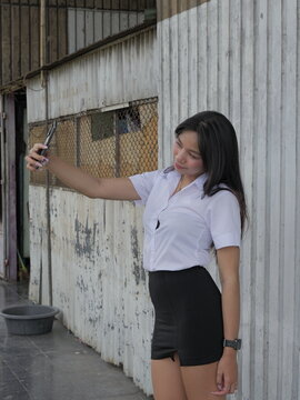 A young Asian woman in a student uniform uses a smartphone to take selfies