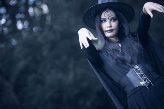 Halloween theme: A young woman with ghostly makeup and hair, dressed as a witch, holding a magic wand and a pumpkin amidst a graveyard; Halloween festival concept.