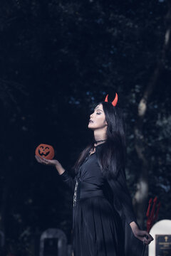 Halloween theme: A young woman with ghostly makeup and hair, dressed as a witch, holding a magic wand and a pumpkin amidst a graveyard; Halloween festival concept.