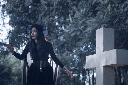 Halloween theme: A young woman with ghostly makeup and hair, dressed as a witch, holding a magic wand and a pumpkin amidst a graveyard; Halloween festival concept.