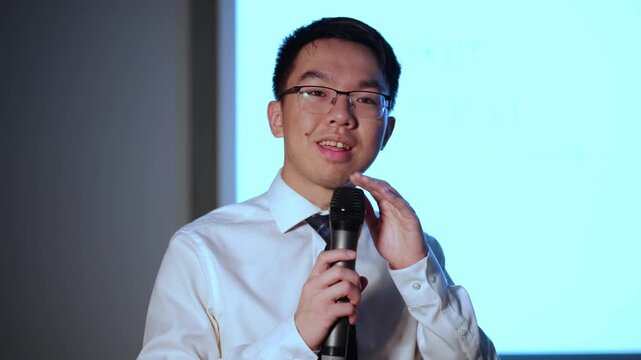 A young Asian man in glasses, a white shirt, and tie taps a handheld microphone during a sound check in front of a projection screen, for audio testing, event preparation, public speaking concepts.