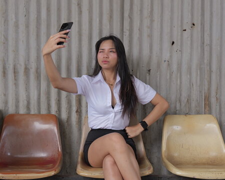 A young Asian woman in a student uniform winks as she takes selfies with a smartphone