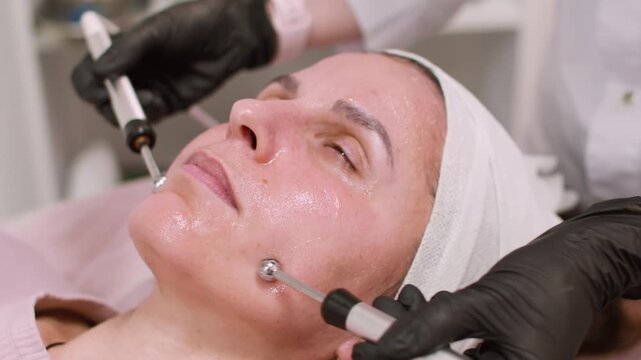 Facial electrotherapy. Female procedure. Esthetician in black gloves using professional modern device for lifting client skin in salon.