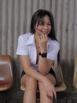 A young Asian female university student sits on a plastic chair, with a corrugated wall in the background