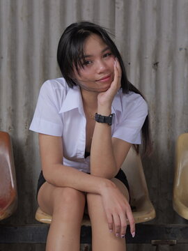 A young Asian female university student sits on a plastic chair, with a corrugated wall in the background