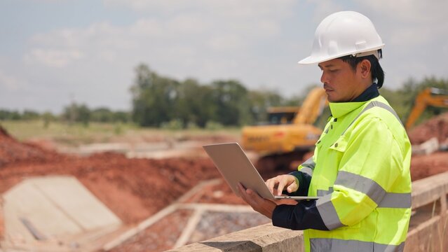 Asian male civil engineer using laptop computer and radio transceiver to supervise bridge construction. Heavy civil infrastructure engineering, technology and site management concept.