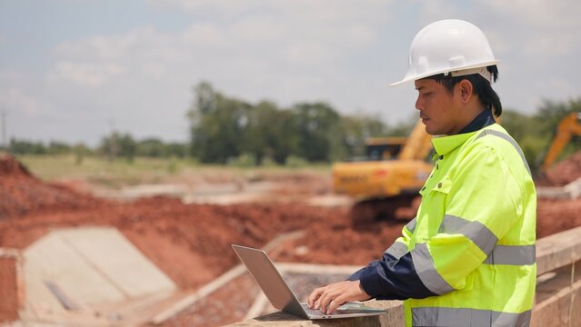 Asian male civil engineer using laptop computer and radio transceiver to supervise bridge construction. Heavy civil infrastructure engineering, technology and site management concept.