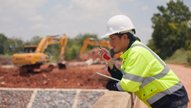 Asian male civil engineer using laptop computer and radio transceiver to supervise bridge construction. Heavy civil infrastructure engineering, technology and site management concept.
