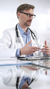 Doctor man, wearing a stethoscope, blue shirt and a lab coat is gesturing with hands overs a glass desk with laptop in clinic office, vertical portrait view. Medicine and science