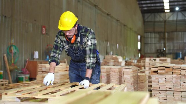 A skilled carpenter wearing protective equipment assembles a wooden pallet among stacked lumber inside an industrial woodworking factory.