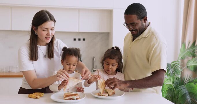 Happy interracial couple making sandwiches together with little kids at home kitchen table teaching daughters basic food preparation skills and simple household chores praising children for helping