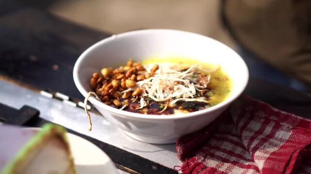 A bowl of traditional Indonesian chicken porridge served in a white ceramic bowl with shredded meat, fried peanuts, and savory yellow broth.