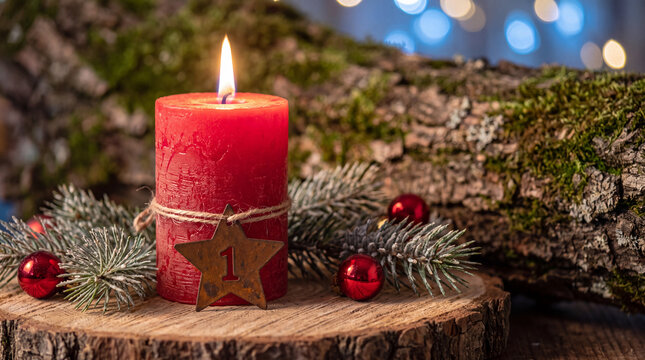 A festive red advent candle with a star-shaped number one tag, surrounded by evergreen branches and holiday ornaments on a rustic wooden slice.
