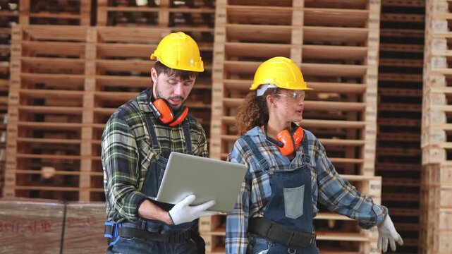 Two industrial workers wearing hard hats and protective gear inspect wooden pallets in a factory while using a laptop for production planning and quality control.
