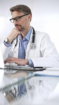 Doctor man in a blue shirt under a medical coat is thinking while using technology in medicine. Portrait of physician typing on a laptop on a glass desk in clinic, vertical view. Medicine and science