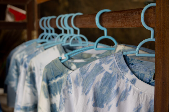 close-up shot of tie-dye t-shirts hanging on light blue plastic hangers. The shirts feature a soft, pastel tie-dye pattern.
