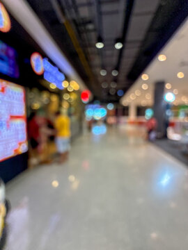Blurred image of people walking down a wide, well-lit corridor in a modern shopping mall or large retail center, with colorful bokeh lights.