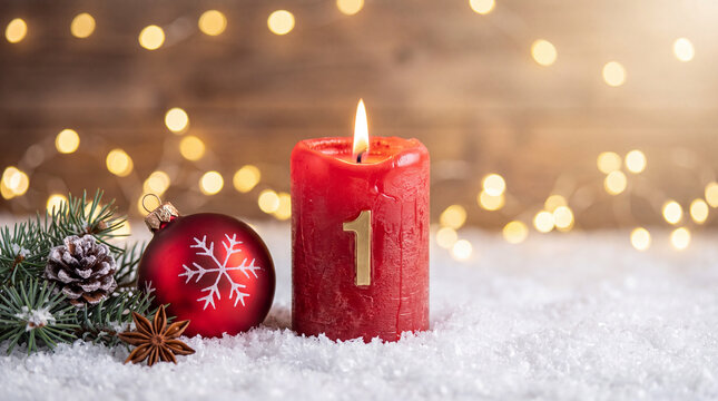 A burning red advent candle with the number one, next to a festive bauble, pine branch, and star anise on snow against a blurred bokeh background.