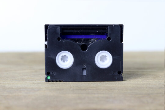 A black MiniDV video cassette tape standing vertically on a wooden surface against a plain light background
