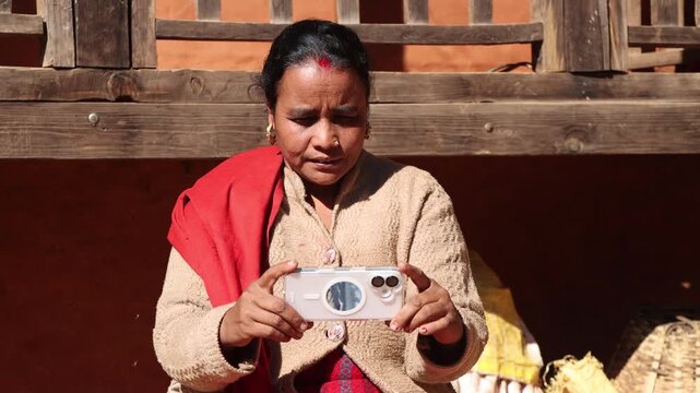 A traditional rural woman is attentively using a modern smartphone outside her village home.